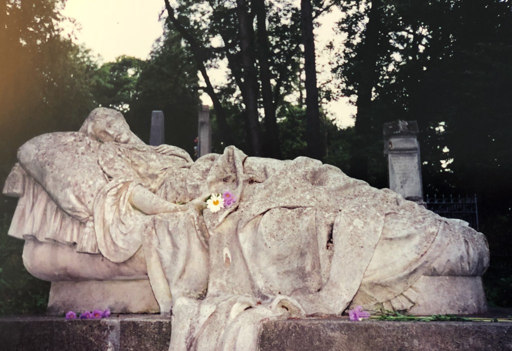 Photograph of a tombstone of a woman lying on a bed surrounded by fresh flowers. From the Lychakiv Cemetery in Lviv.