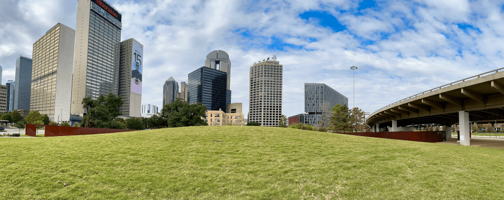 Photograph: A ribbon of COR-TEN steel slices through a grassy hillock in a park in downtown Dallas, Texas. On the horizon sit several skyscrapers. On the right, a raised highway swerves out of frame.